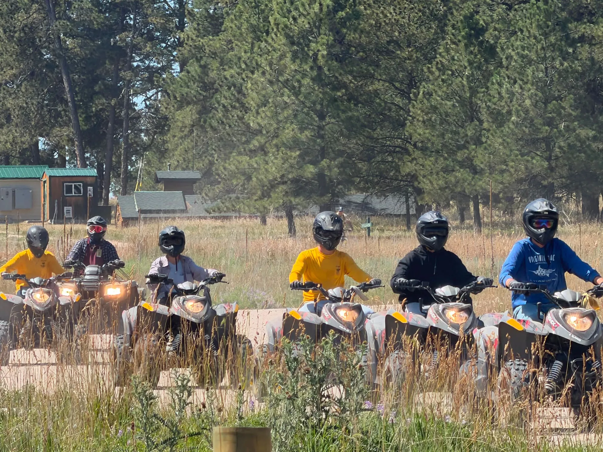 Scouts wearing helmets sit on all-terrain vehicles in a grassy pine clearing.