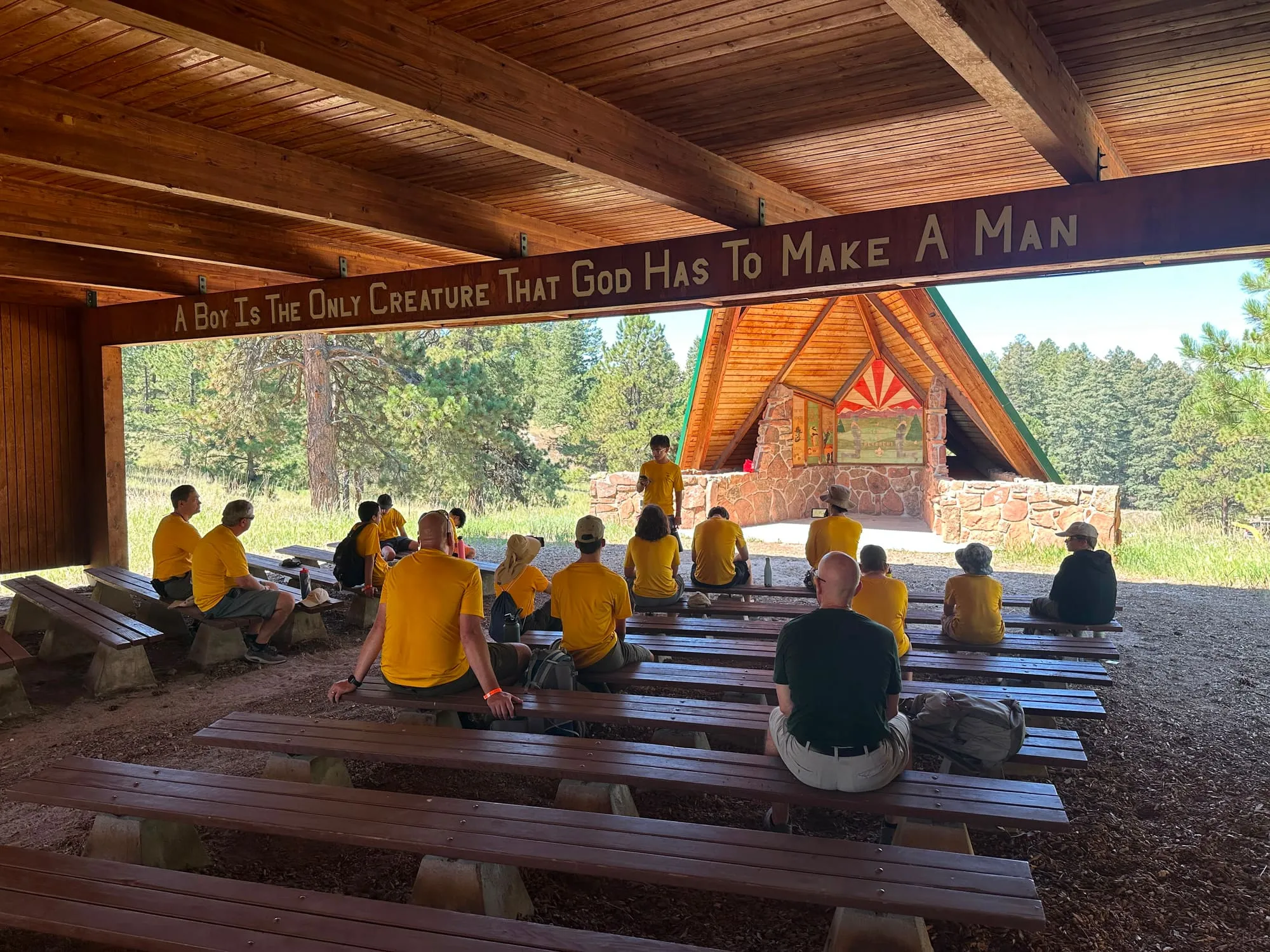 Scouts and adult leaders sit on benches under a wooden pavilion, facing a camp chapel.
