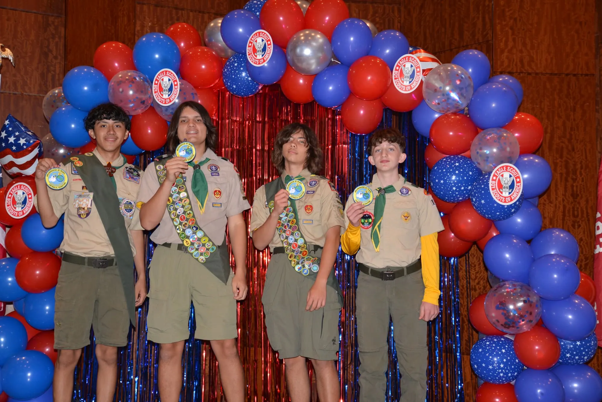 Four scouts in medal-covered sashes hold Eagle medals in front of a red, white, and blue balloon arch.