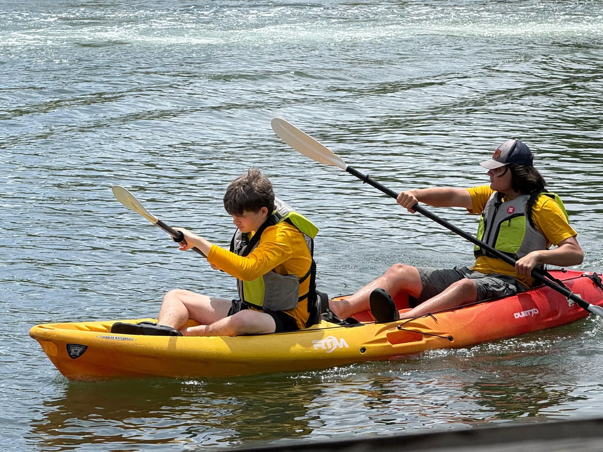 Two scouts in life jackets paddle a yellow and orange kayak.