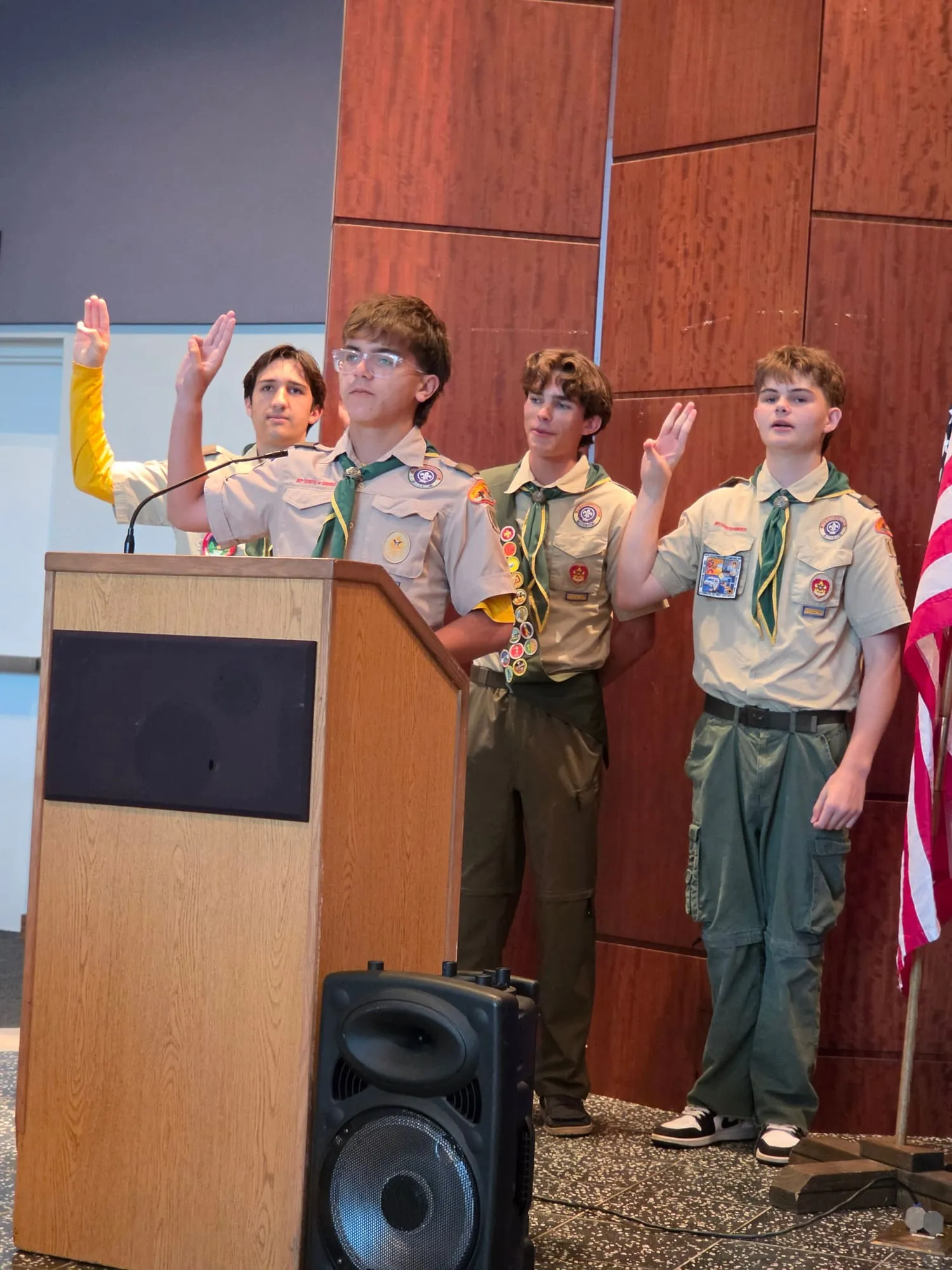 Four scouts in uniform stand at a wooden lectern and raise their hands during a ceremony.