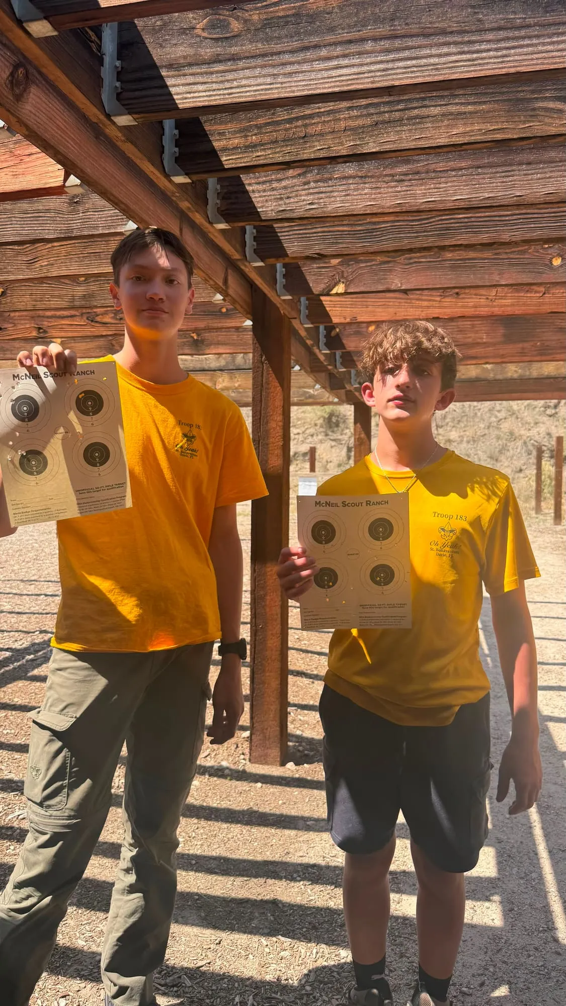 Two scouts hold up rifle targets under a wooden range shelter at summer camp.