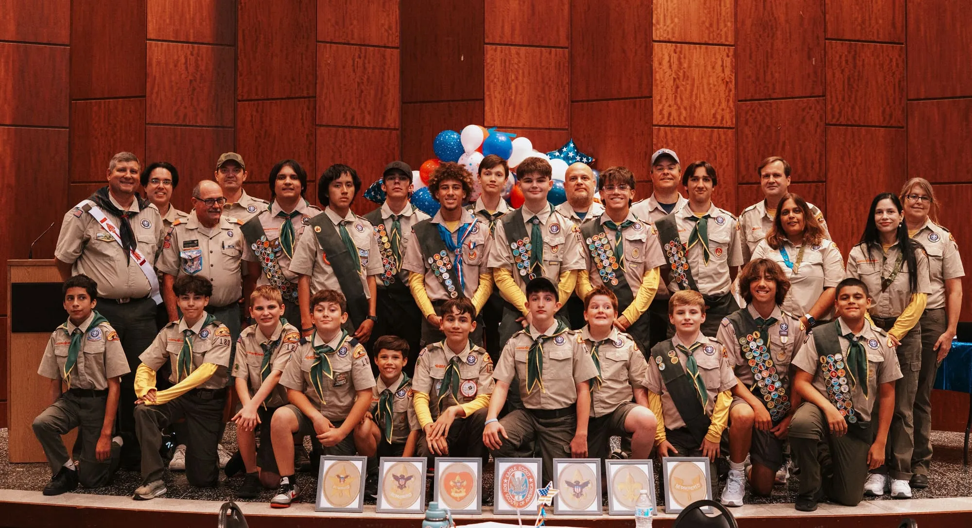 The troop posed together on stage at a Court of Honor, in tan uniforms, with award plaques lined up in front.